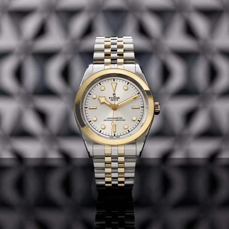 A M79683-0002 wristwatch with a silver and gold band, and a white dial with gold accents, is displayed against a geometric patterned background.