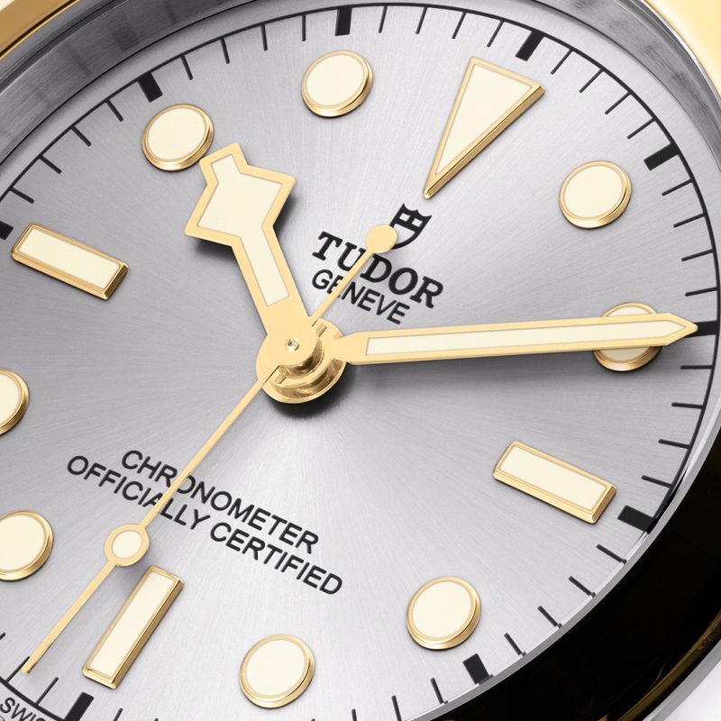 Close-up of a Tudor M79603-0002 watch dial with gold hour, minute, and second hands, marked with "Chronometer Officially Certified" and the brand's logo. The dial features geometric hour markers on a silver background.