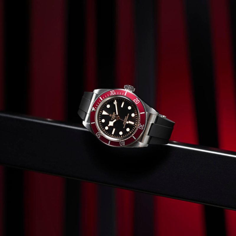 A close-up of a wristwatch with a black dial, red bezel, and black rubber strap, displayed against a dark background with red and black vertical stripes. M7941A1A0RU-0002