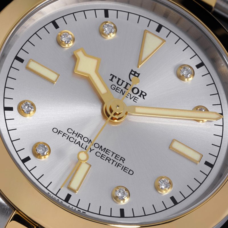 Close-up of a Tudor Geneve wristwatch face, displaying "CHRONOMETER OFFICIALLY CERTIFIED" text. The watch has a silver dial, gold markers, gold hands, and diamond accents at each hour mark.