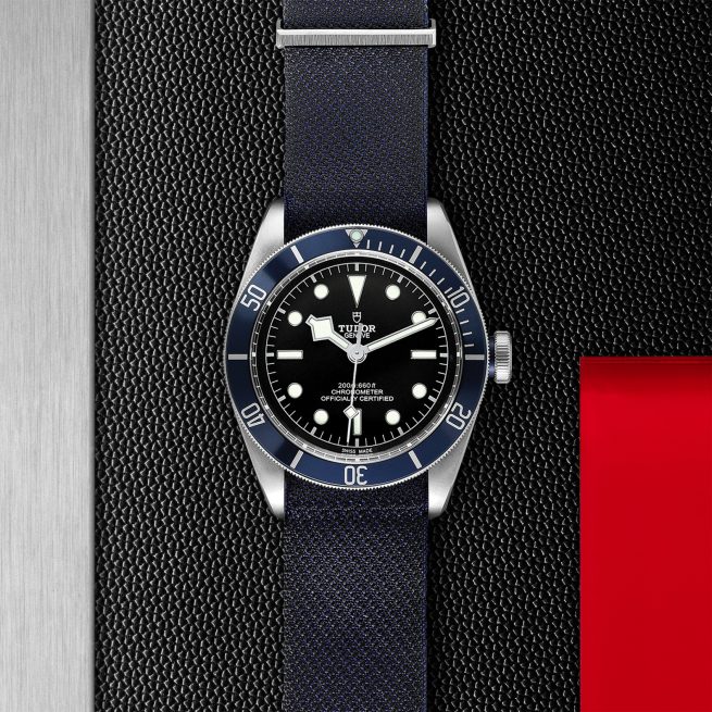 A Tudor M79230B-0006 wristwatch, featuring a black dial and bezel with silver markers and hands, paired with a black fabric strap, is positioned on a textured black surface next to a metal and red section.