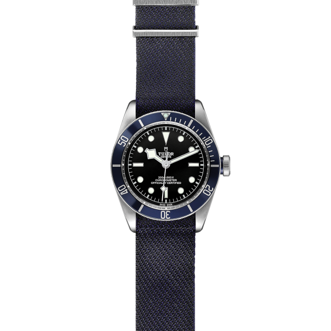 The M79230B-0006 wristwatch features a black dial with white markers, a blue fabric strap, and a blue bezel adorned with white numbers and markers. The brand name "Tudor" is prominently displayed on the dial.