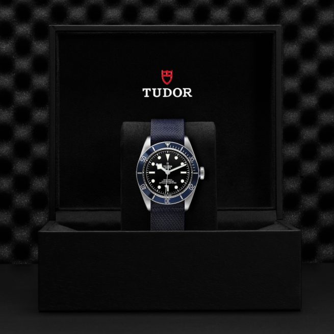 A Tudor wristwatch, model M79230B-0006, with a blue strap is displayed in an open black box with the Tudor logo inside the box lid.