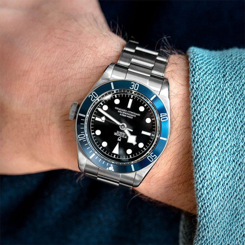 A close-up of a wrist wearing a M79230B-0008 with a blue bezel and black dial, displaying the time as 10:10. The person is dressed in a blue garment.