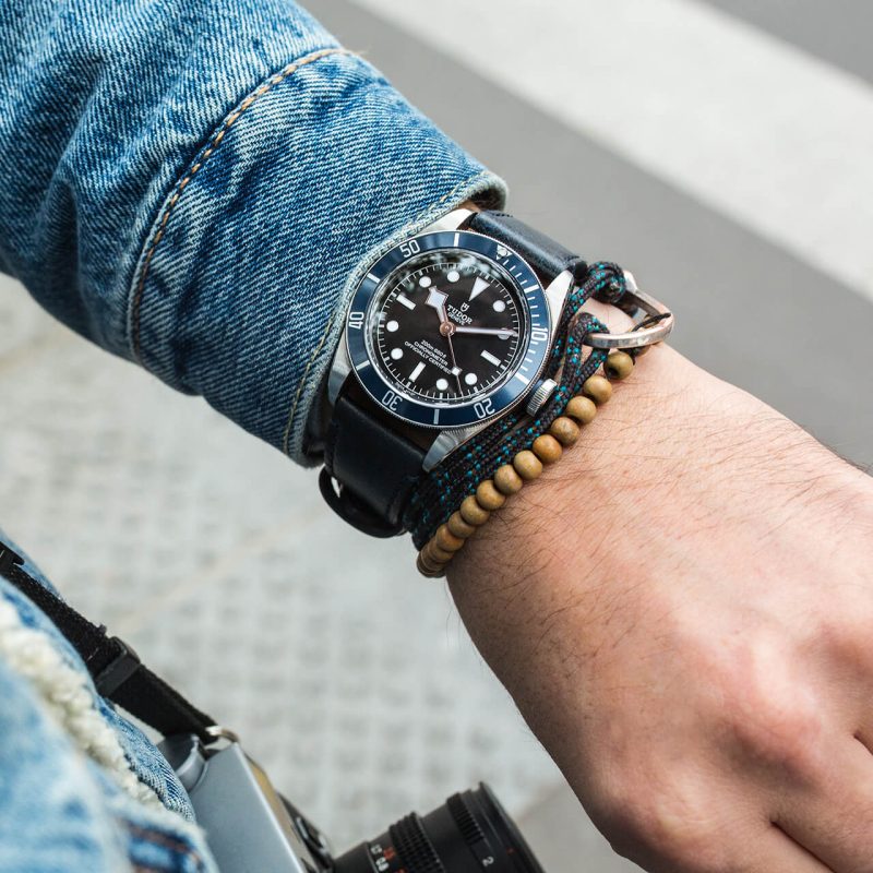 A wrist with a M79230B-0006 and wooden bead bracelet, wearing a denim jacket and holding a camera.