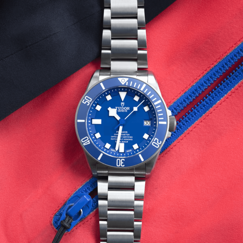 A M25600TB-0001 watch with a blue dial and stainless steel bracelet is placed on a red and black fabric background with a blue zip.