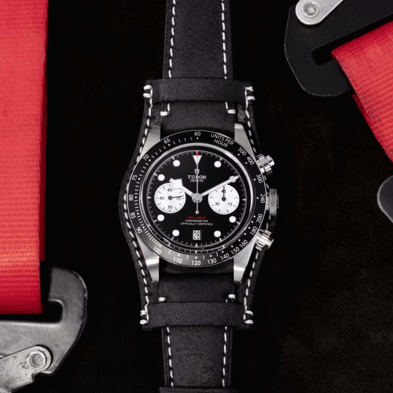 A Tudor M79360N-0005 with a black leather strap is displayed against black and red elements. The watch features a black dial, white subdials, and a tachymeter scale on the bezel.