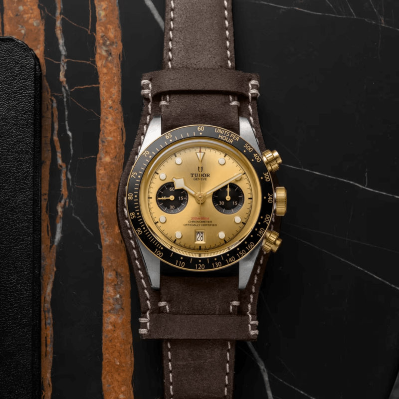 A M79363N-0008 wristwatch with a gold dial and brown leather strap, placed on a dark marble surface with light brown veins.