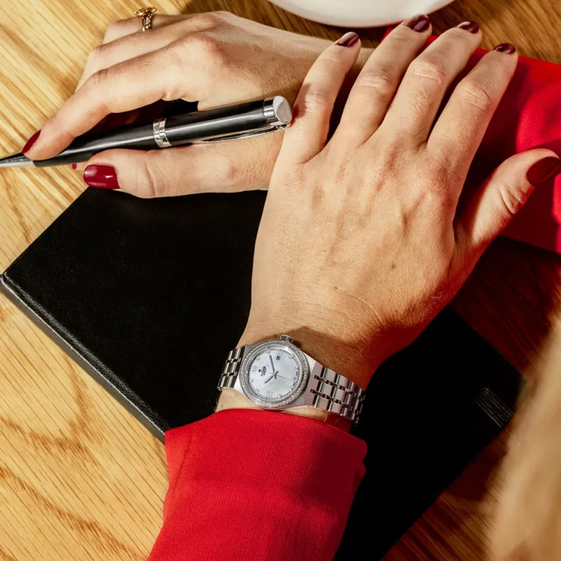 A woman's wrist with a M28320-0001 watch and a pen on a table.