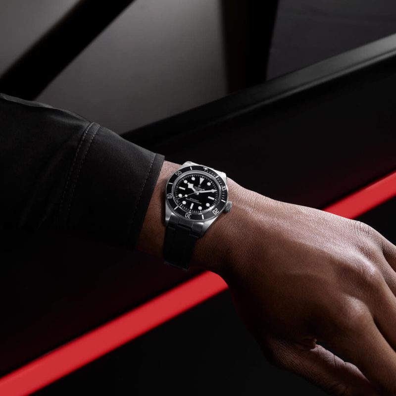 A wrist wearing a sleek, black analog M7941A1A0NU-0002 with a silver bezel and a black strap, against a background featuring red and black elements.