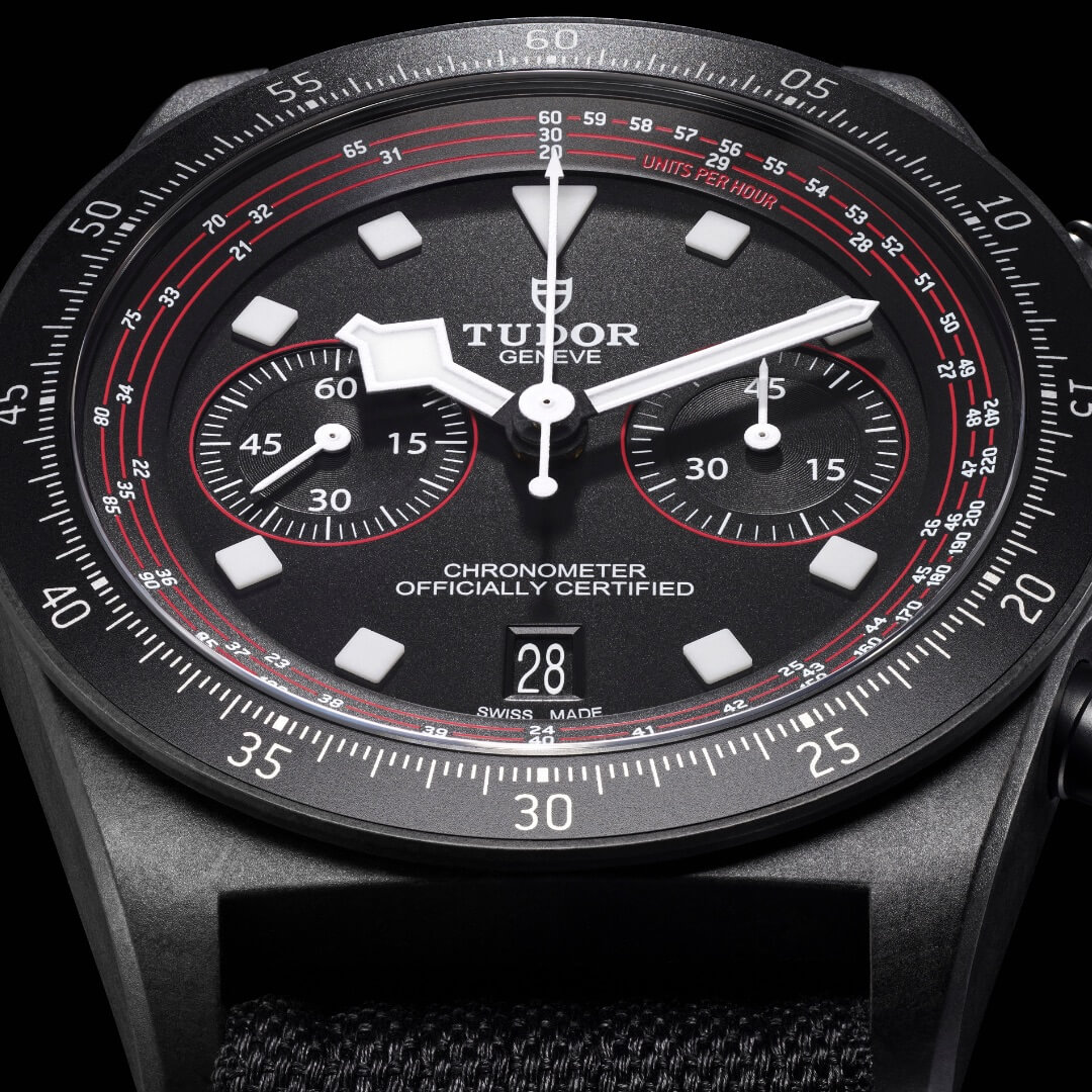 Close-up view of an M25827KN-0001 wristwatch with a black dial displaying chronograph features, including sub-dials for seconds and minutes, date window at 6 o'clock, and red accents on the black bezel.