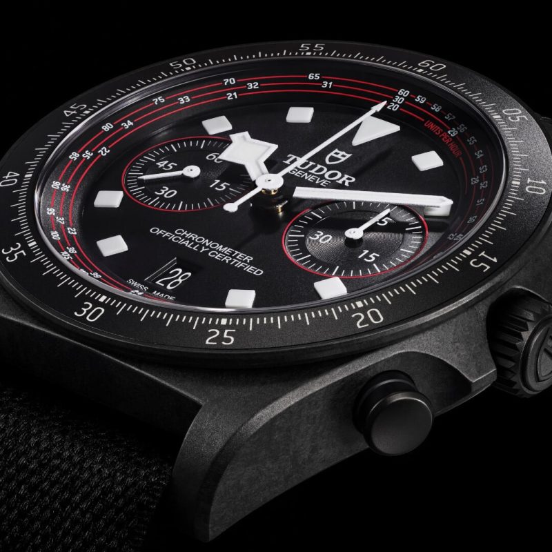 A close-up of a black M25827KN-0001 chronograph watch with a textured strap, three sub-dials, red accents, and a date window displaying "28.