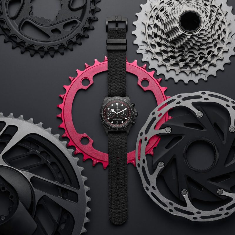 A black wristwatch with a fabric strap is placed among various bicycle gears and cogs on a dark surface.