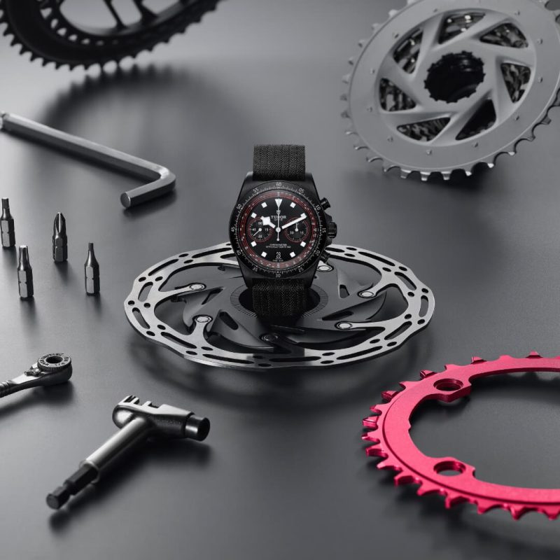 A M25827KN-0001 with a red and black face rests on a bicycle disc brake rotor, surrounded by various bike parts and tools on a black surface.