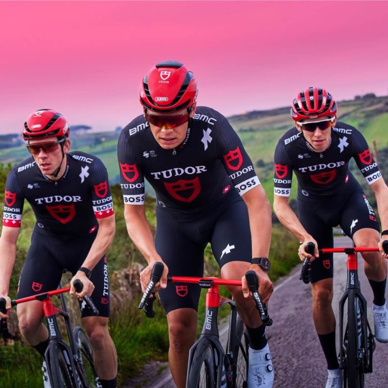 Three cyclists in black and red M25827KN-0001 uniforms ride along a rural path at sunset, with a pink sky and green landscape in the background.