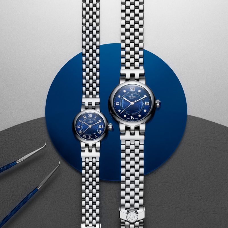 Two silver wristwatches with blue dials are displayed side by side on a dark grey and blue background. Tweezers are positioned next to the watches.