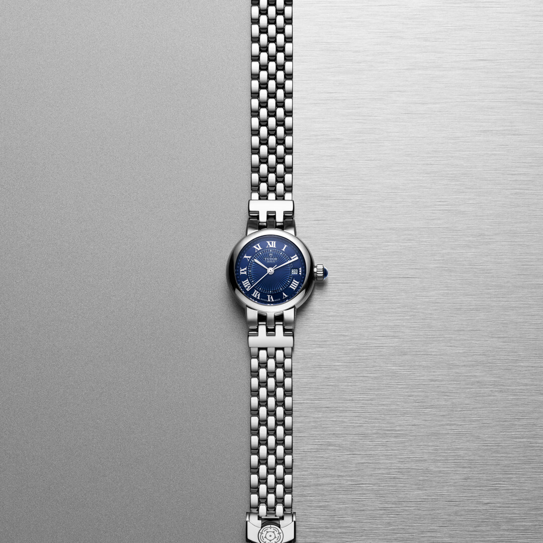 A M35800-0010 with a blue dial and Roman numeral markers, featuring a linked metal band, is displayed against a grey and silver gradient background.