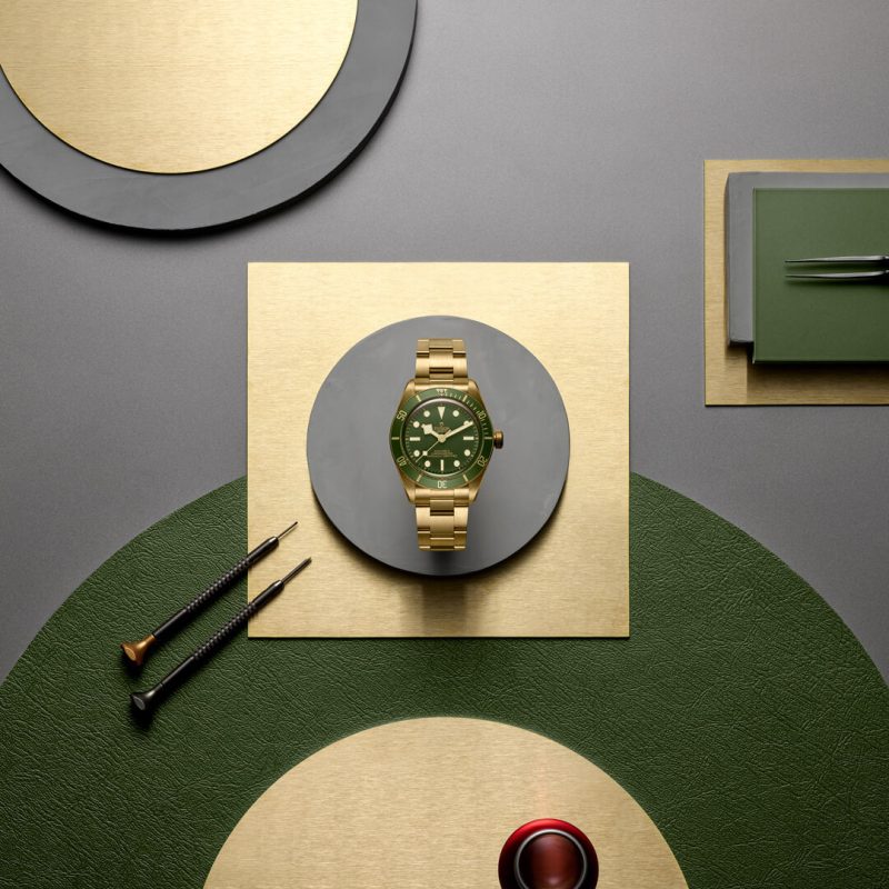 Gold wristwatch with a green dial positioned on circular and square gold and green surfaces, surrounded by watchmaking tools.