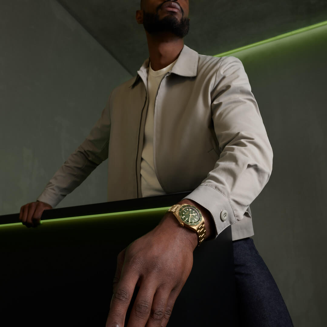 A person wearing a beige jacket and a gold M79018V-0006 stands with one hand resting on a black surface and a green light accent in the background.