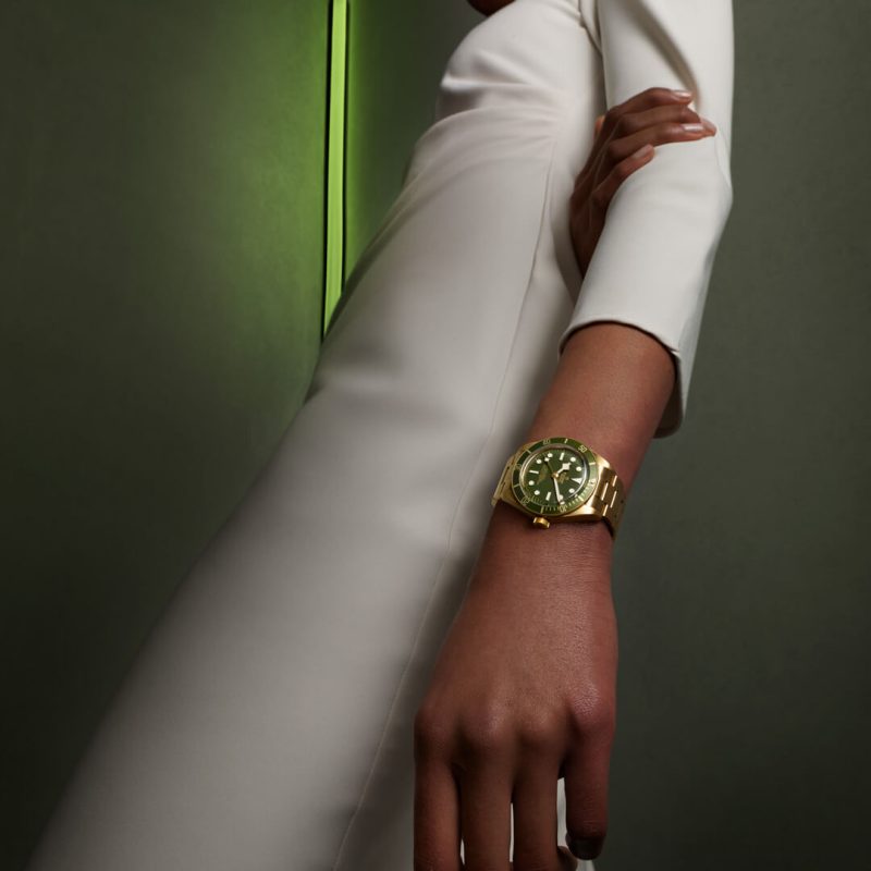 A person wearing a white long-sleeved garment shows an M79018V-0006 with a green watch face against a green background.