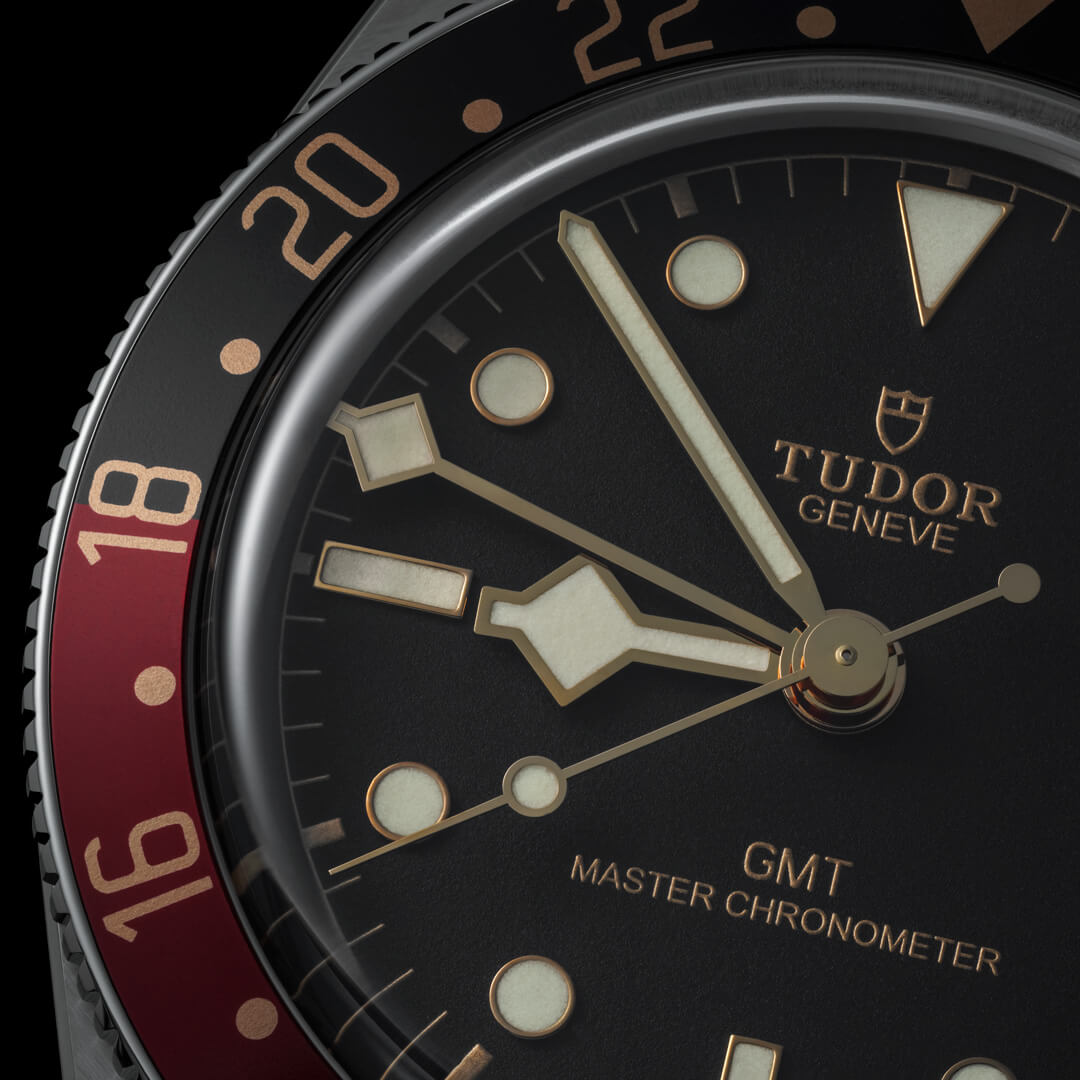 Close-up of a M7939G1A0NRU-0001 wristwatch with a black dial, red and black bezel, and gold-toned markers and hands.