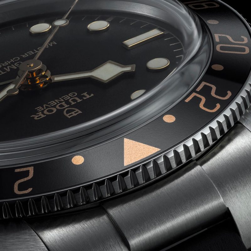 Close-up of a wristwatch featuring a black dial with luminescent hands and markers, a domed crystal, and a stainless steel bracelet. The watch displays the product name "M7939G1A0NRU-0001" on the dial.