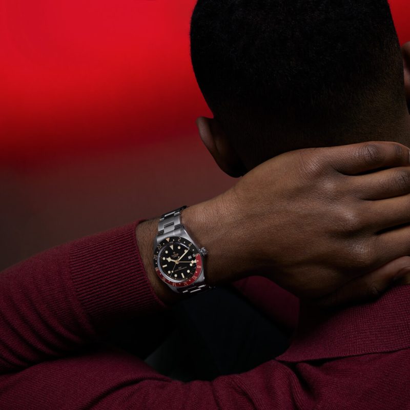 A person with dark hair wearing a M7939G1A0NRU-0001 with a black dial and red accents, sitting against a red and dark background. Their hand is resting on the back of their neck.