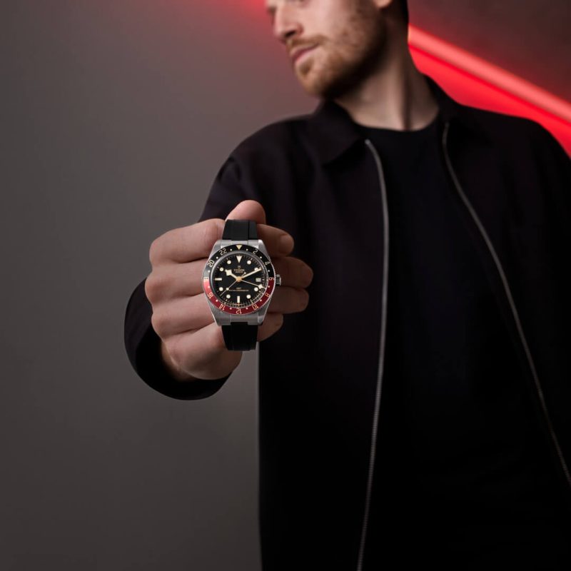 A person in a black jacket holds out a M7939G1A0NRU-0001 with a red and black bezel against a dark background illuminated by a red light streak.