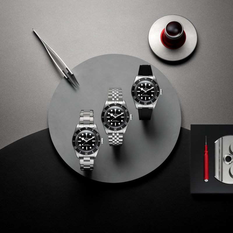Three luxury wristwatches with black dials are displayed on a round, gray surface with a pair of tweezers, a loupe, and a red-handled tool nearby.