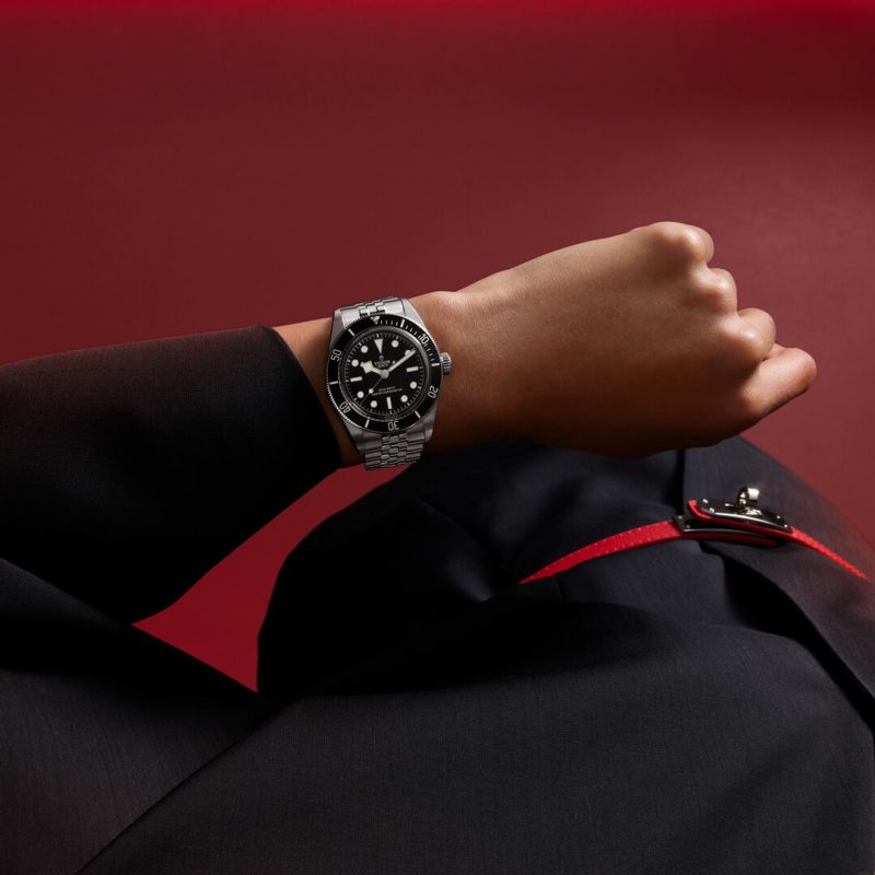 Close-up of a person wearing a black suit and a silver M7941A1A0NU-0002, with the wrist positioned in front of a dark red background.