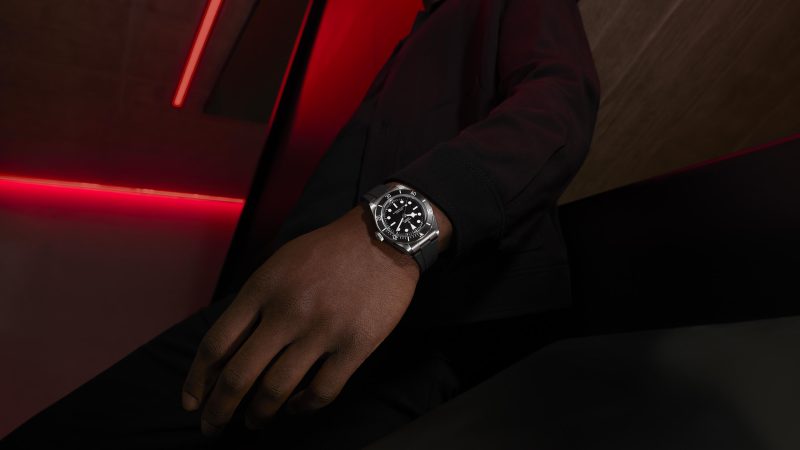 A person wearing a black jacket shows a close-up of their wrist with a silver wristwatch, against a background illuminated by red and black lighting.