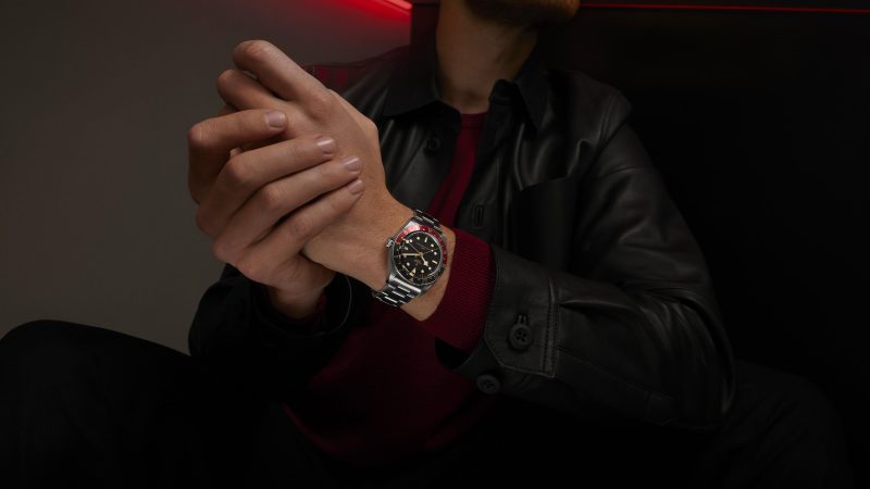 A person wearing a black leather jacket and a red shirt is clasping their hands, displaying a wristwatch with a metal band and a dark face.