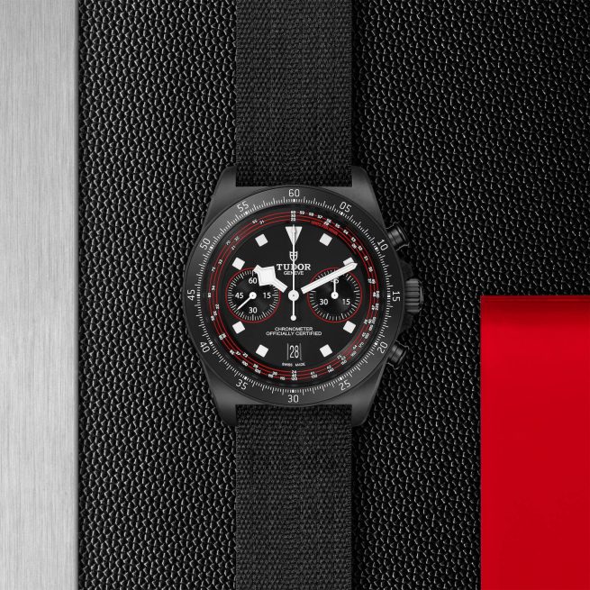 A black M25827KN-0001 wristwatch with a red and white bezel, black fabric strap, and chronograph dials, displayed against a metallic and red background.