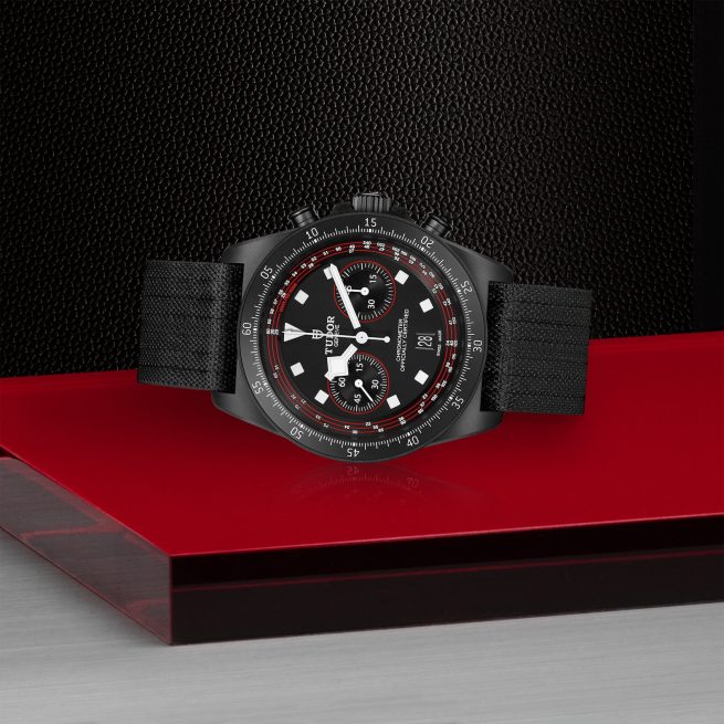 A M25827KN-0001 with a black fabric strap, red and white accents on the dial, and a tachymeter bezel is displayed on a red and black surface.