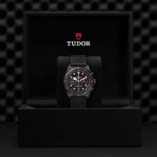 A M25827KN-0001 watch with a black dial and red accents is displayed in an open black box with the Tudor logo on the lid. The box interior is lined with black material.