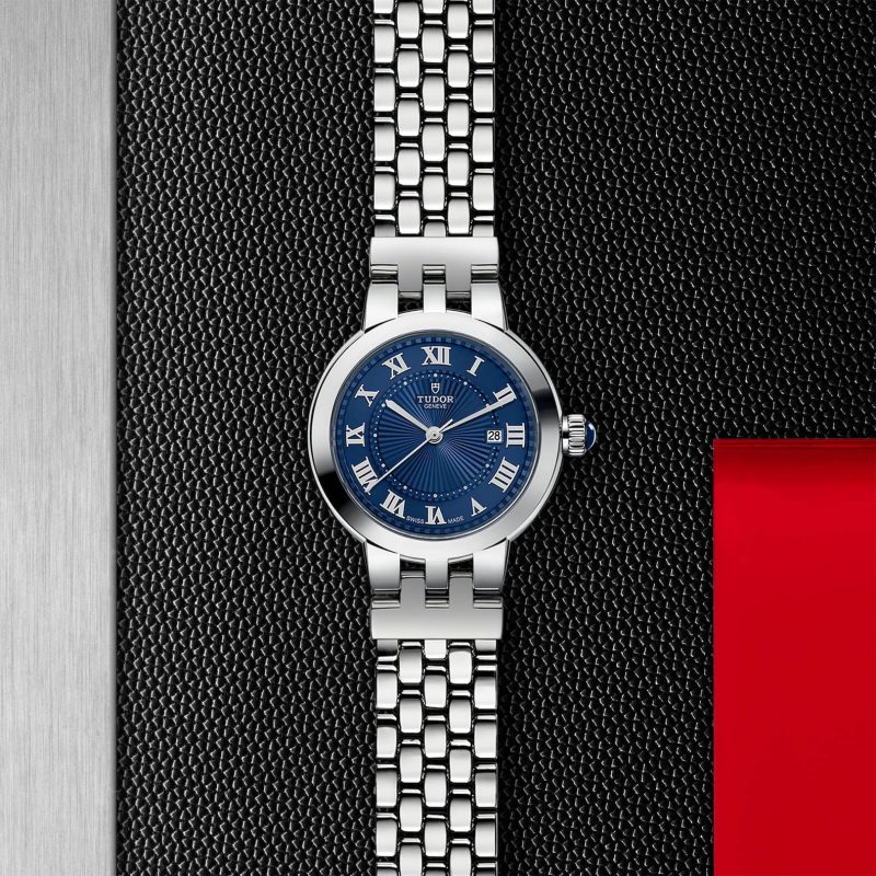 Silver wristwatch with a blue dial, featuring Roman numerals for hour markers, on a textured black and red background.