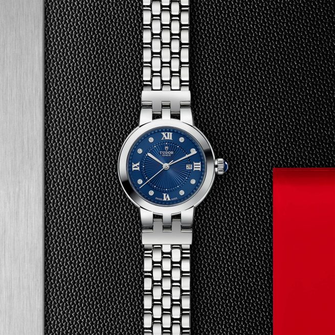 A silver wristwatch with a blue face featuring Roman numerals and a date display sits on a textured dark background with a red accent on the right.