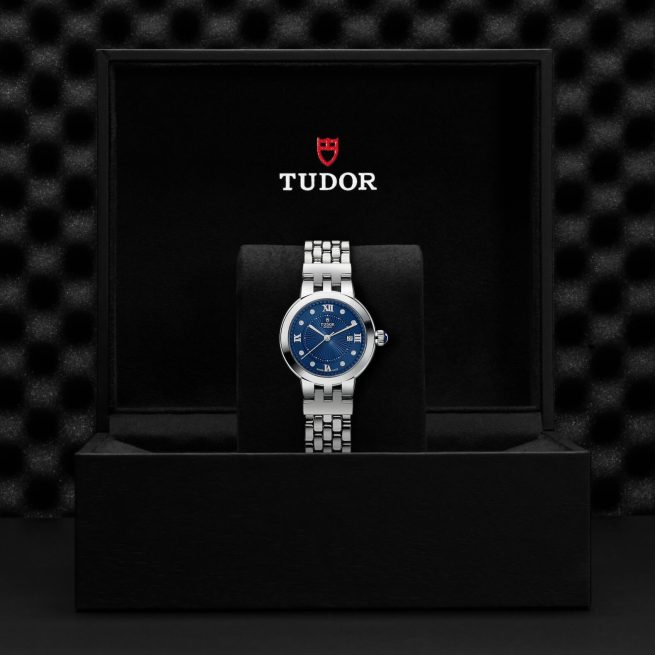 A silver Tudor wristwatch with a blue dial in a black presentation box. The box has the Tudor logo and name printed in white and red.