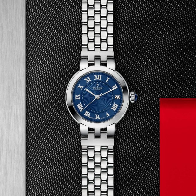 A silver wristwatch with a blue dial featuring Roman numerals and a date display on a metal mesh background.