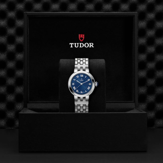 A stainless steel wristwatch with a blue dial and Roman numerals is displayed in a black box. The box features the brand name "Tudor" and its logo.