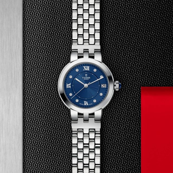 A silver wristwatch with a blue dial, Roman numerals, and a metal bracelet displayed on a black textured background with a red accent on the right.