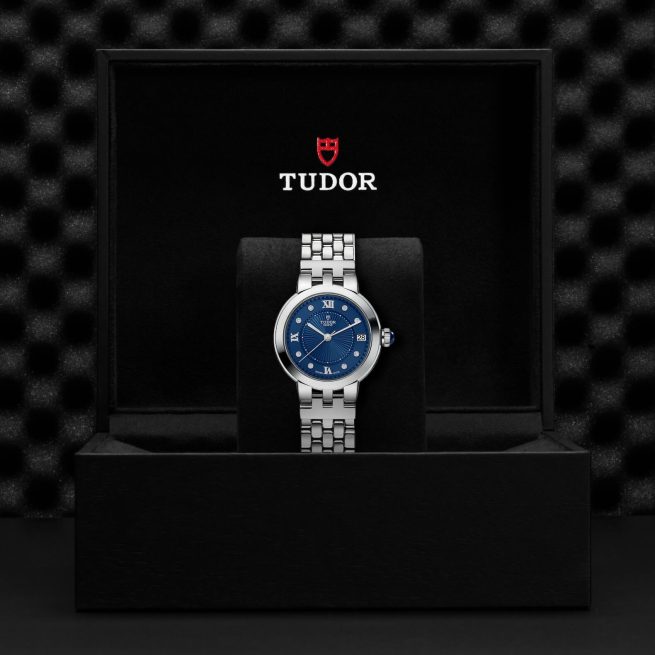 A luxury wristwatch with a blue dial and silver strap is displayed in an open black Tudor-branded box.