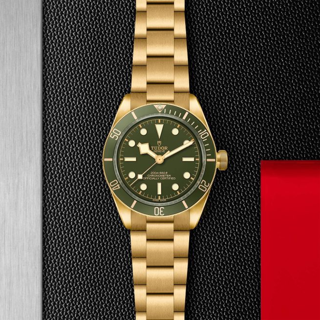 A gold wristwatch with a green face and bezel, featuring a gold bracelet, is displayed against a textured black background with a red accent.