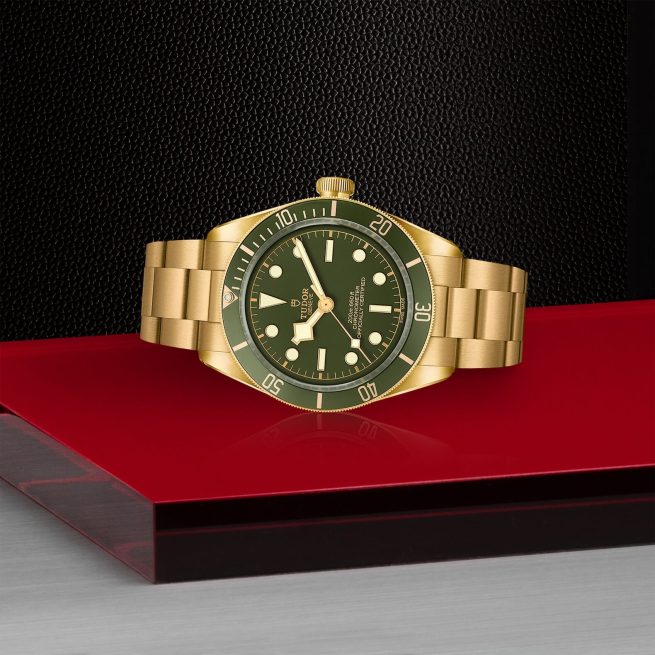 A gold wristwatch with a green dial and bezel, featuring luminous markers and a stainless steel bracelet, displayed on a red surface against a black textured background.