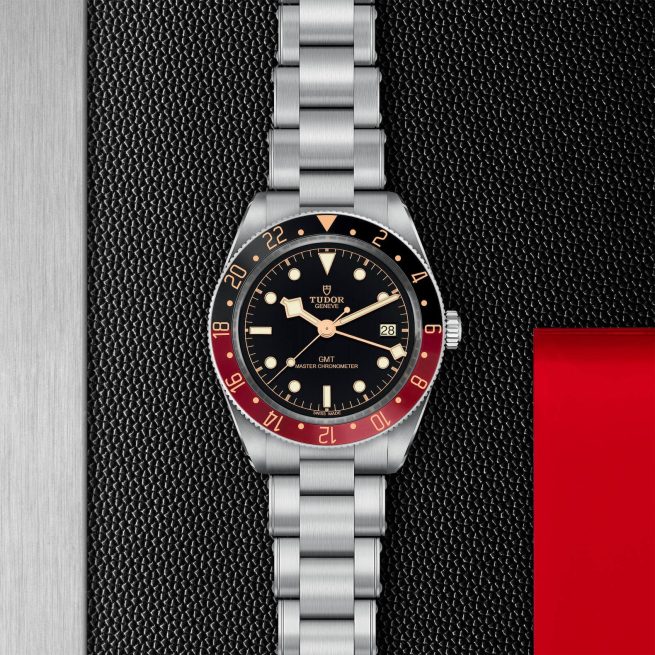 A M7939G1A0NRU-0001 wristwatch with a stainless steel strap, black dial, and a red and black bezel is displayed on a textured black background.