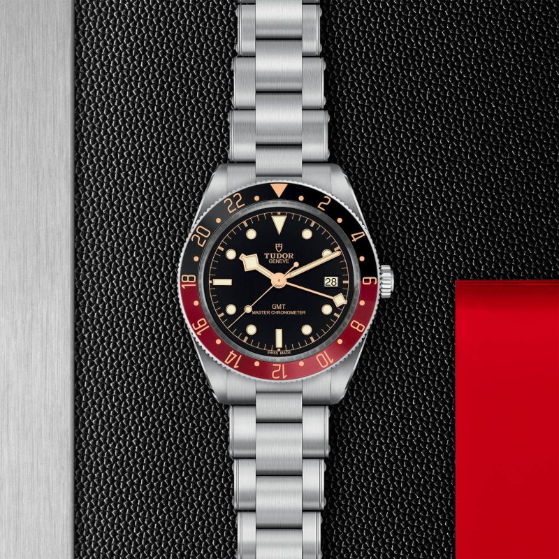 A M7939G1A0NRU-0001 wristwatch with a stainless steel strap, black dial, and a red and black bezel is displayed on a textured black background.