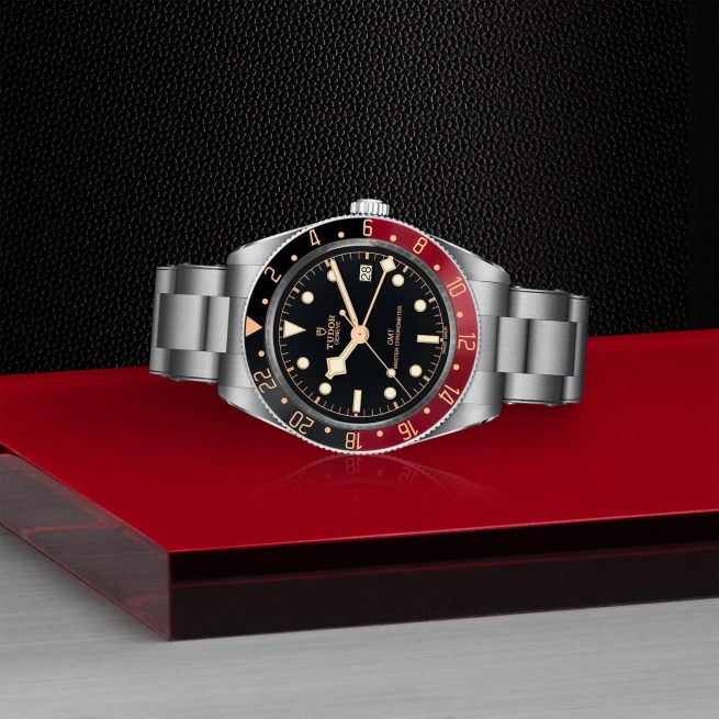 A M7939G1A0NRU-0001 with a black dial, red and black bezel, and metal strap displayed on a red and black surface.