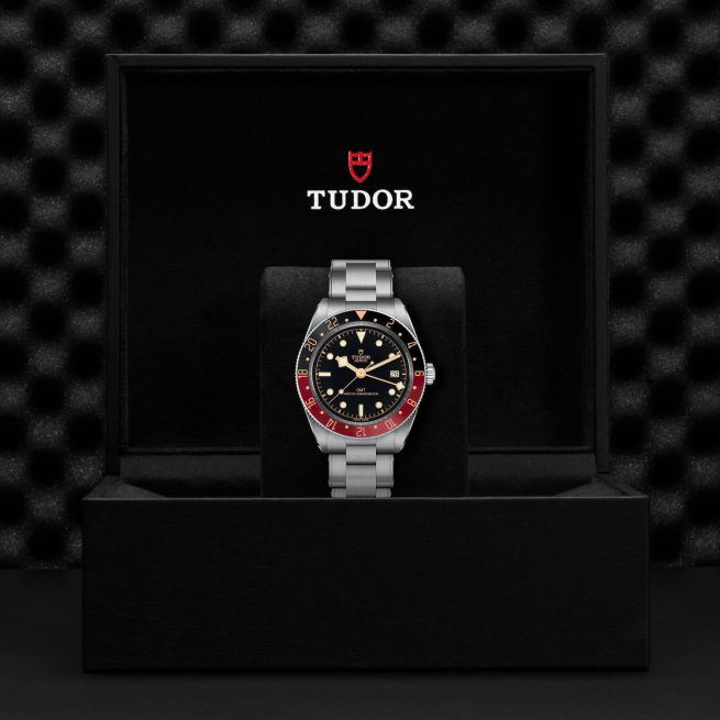 A M7939G1A0NRU-0001 wristwatch with a stainless steel band, black dial, and red and black bezel is displayed in an open black box with the Tudor logo on the inside lid.