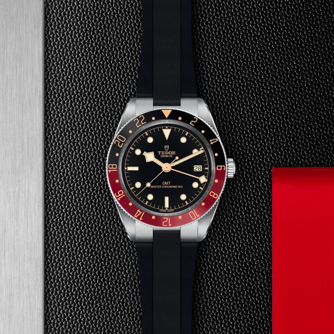 A M7939G1A0NRU-0002 wristwatch with a black and red bezel, black dial, and black strap is positioned against a textured, patterned background.