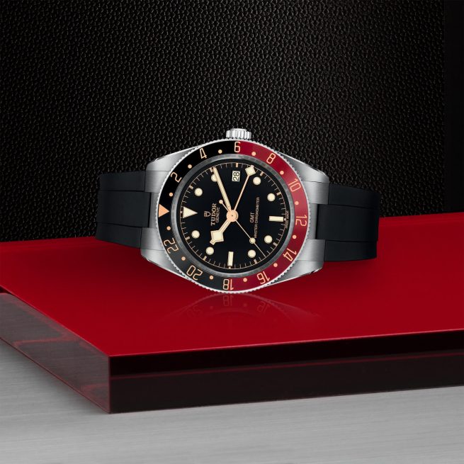 A M7939G1A0NRU-0002 with a black dial, red and black bezel, silver case, and black strap is displayed on a red surface with a dark background.